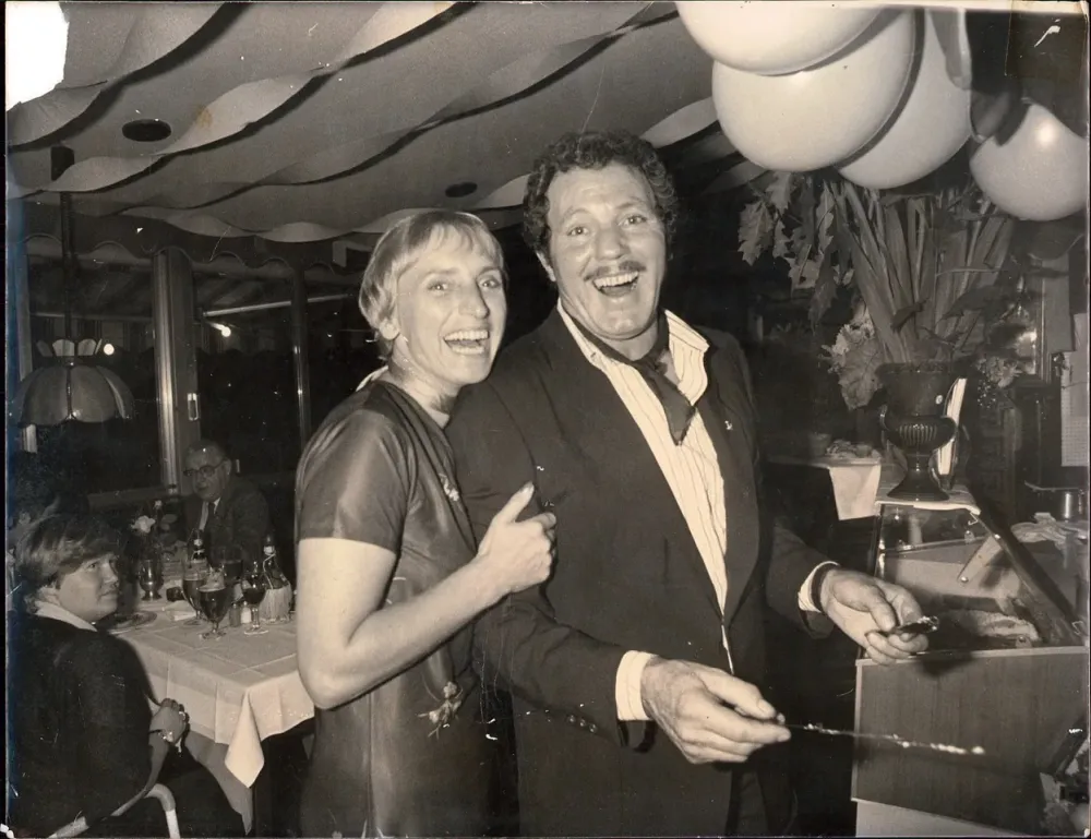 Schwarz-Weiß-Foto: Franco Francucci senior lachend mit einem Gast am Buffet seines Lokals, Luftballons unter der Decke