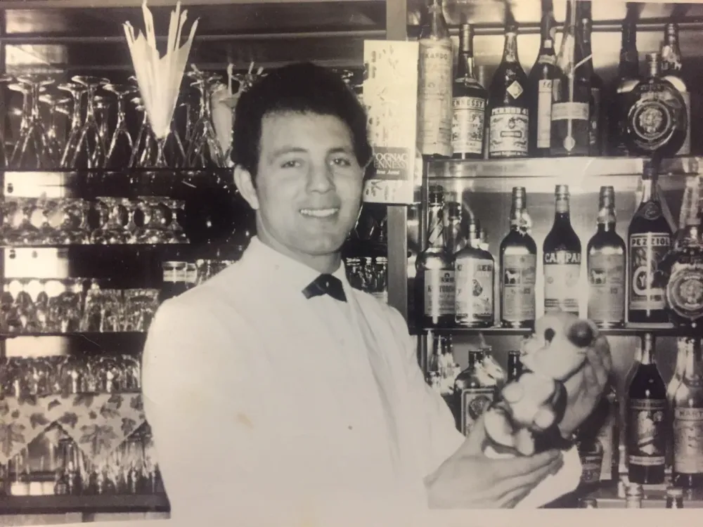Schwarz-Weiß-Foto: der junge Franco Francucci senior in weißer Kellnerjacke und Fliege hinter einer Bar