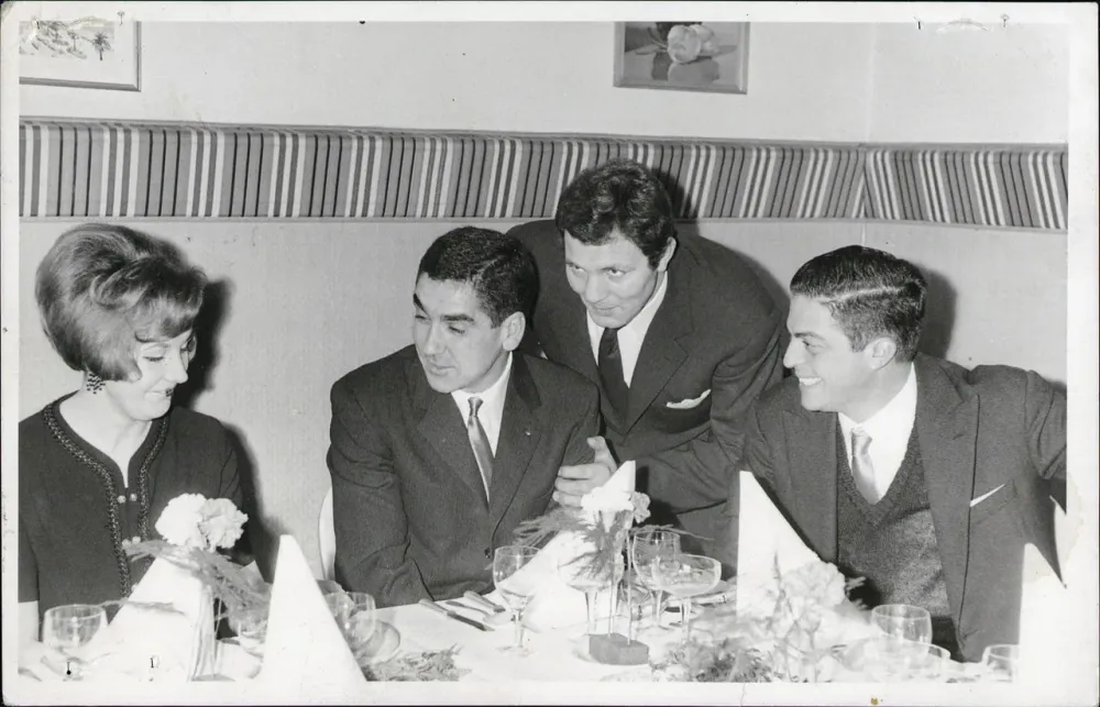 Schwarz-Weiß-Foto: Franco Francucci senior beugt sich lächelnd zu drei Gästen an einer festlich gedeckten Tafel